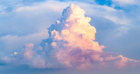Towering cloud formation rises against a soft blue sky, glowing with warm light that reveals dramatic volume, gentle color transitions, and the calm beauty of changing atmospheric conditions.
