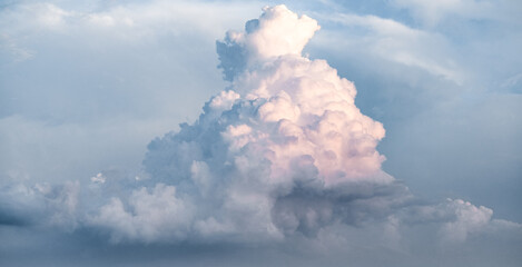 Towering cloud formation rises against a soft blue sky, glowing with warm light that reveals dramatic volume, gentle color transitions, and the calm beauty of changing atmospheric conditions.