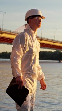 Young environment inspector in hazmat suit and hard hat stands by river and a busy bridge at sunset, clipboard in hand, observing water quality, collecting samples and checking for pollution, vertical