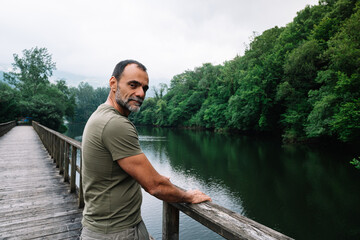 A man stands on a bridge overlooking a river. The man is wearing a green shirt and has a beard. The scene is peaceful and serene, with the man looking out over the water
