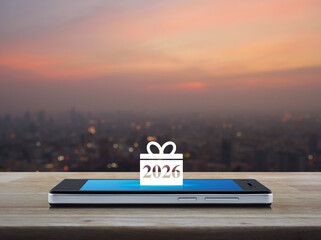 Gift box happy new year 2026 flat icon on modern smart mobile phone screen on wooden table over blur of cityscape on warm light sundown, Business happy new year 2026 shop online concept
