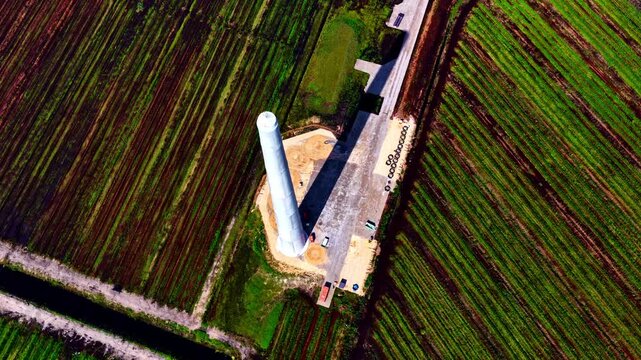 A tall white wind-turbine tower section stands on a construction pad surrounded by green striped fields, viewed directly from above as crews prepare the site for further assembly.