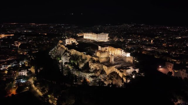 Panoramic night drone orbit capturing the city of Athens with the illuminated Acropolis rock at the center, surrounded by glowing urban lights and landmarks.