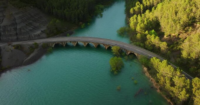 Drone tracking cyclists across arched road bridge over turquoise waters near Antiguo Balneario de Tiermas, Embalse de Yesa Reservoir, Aragon, on Camino de Santiago corridor area, Spain