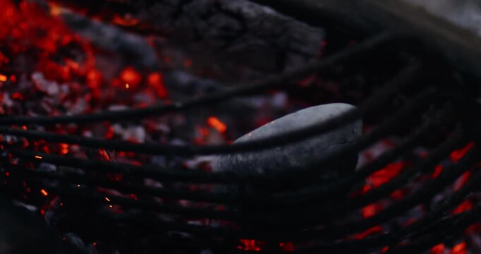 Extreme close-up lingers on glowing red embers while a pale, ash-covered stone rests at the edge of the frame, with every spark and shift in the coals slowed down for maximum texture.