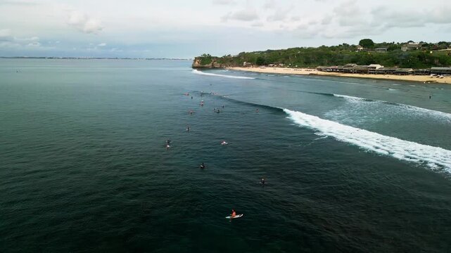 Drone video capturing a wide surf lineup with multiple surfers floating in deep water as smooth wave sets roll toward a sandy shoreline along a tropical Bali coastal environment