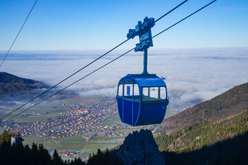 A blue cabin of the Kampenwand cable car hovers above Aschau in Bavaria, offering breathtaking views over a sea of fog above Lake Chiemsee
