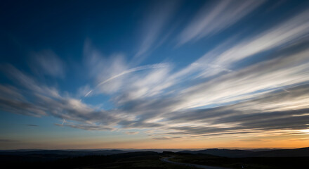 Long exposure photograph capturing the dramatic streaky clouds across a vast sky during either sunrise or sunset, with a dark horizon.