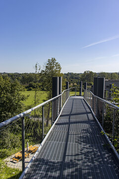 Modern&iacute; ocelov&aacute; l&aacute;vka s kovov&yacute;m ro&scaron;tem v krajině. Perspektivn&iacute; pohled na vyhl&iacute;dkovou cestu s kovov&yacute;m z&aacute;bradl&iacute;m, obklopenou zelen&iacute; pod modrou oblohou. Architektonick&yacute; detail.