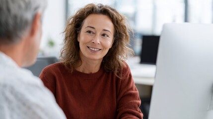 Middle-aged woman with curly hair sitting in front of a computer monitor in an office. she is wearing a red sweater and is smiling at the camera.