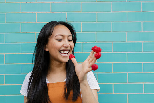 Smiling woman playfully eating raspberries from fingers in front of blue wall