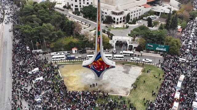 An aerial view of thousands of Syrians celebrating the end of the war in Damascus, carrying Syrian flags.