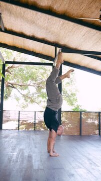 Strong bald man with tattoos practicing an advanced handstand variation in a beautiful, bright yoga shala surrounded by tropical nature. Vertical