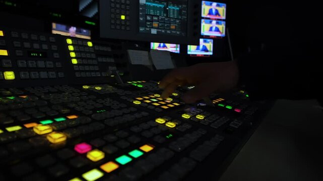 Dimly lit television studio hands operate a professional video switcher console with glowing buttons during a live broadcast production, capturing precise control and focus