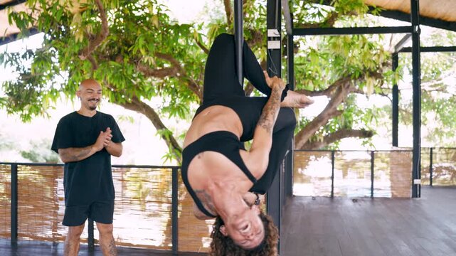 Athletic woman with curly hair mastering a difficult pole dance inversion while her male coach assists and applauds in an outdoor studio