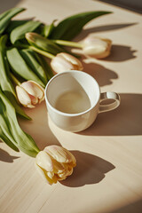 A delicate white coffee cup adorned with intricate handle details and a subtle sheen, filled with a warm brown liquid, sits elegantly beside a bouquet of fresh tulips in various shades of pink 