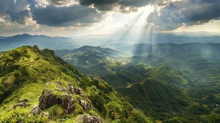 Obraz premium A vibrant green mountain ridge dominates the foreground as powerful sun rays descend from cloudy skies onto the distant, hazy valleys below.