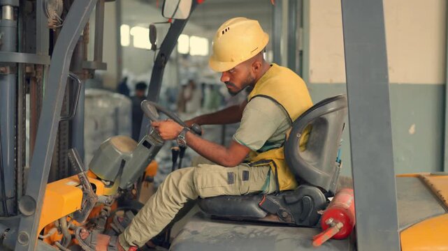 Dynamic handheld orbital shot of a fit male worker wearing a yellow safety vest and helmet operating a grey and yellow forklift inside a busy tent factory in Sialkot.