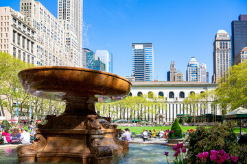 A vibrant, sunlit photo captures people relaxing in New York City&rsquo;s Bryant Park, featuring the iconic Josephine Shaw Lowell Memorial Fountain in the foreground against a backdrop of historic architect