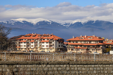 Modern residential buildings with terracotta roofs, set against the majestic snow-covered Pirin mountain range on a crisp, clear winter day in Bansko, Bulgaria
