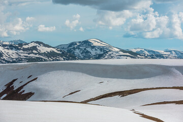 Scenic alpine landscape with sunlit snowfield with snow cornice above abyss against large snow-capped rock mountain top. Sunlight and shadows on snowy field near rocky precipice edge in high mountains © Daniil