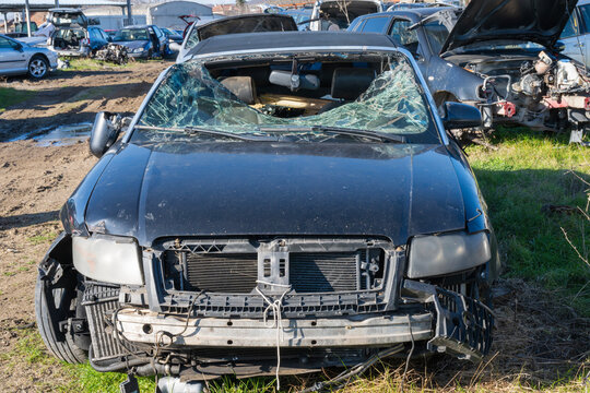 a black convertible with a completely shattered windshield and missing front bumper, parked on a grassy patch in a mud-strewn salvage yard.