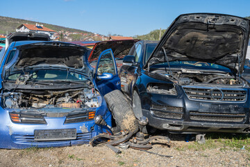 a wrecked blue car and a black SUV parked side-by-side at a salvage yard with their hoods propped open, revealing stripped engine bays and scattered suspension parts on the grassy ground. © Andrey