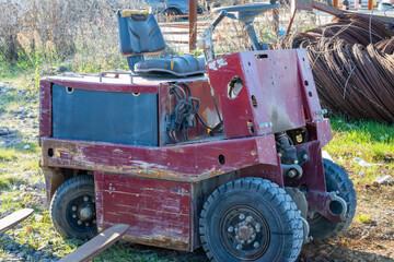 a weathered red industrial tow tractor or utility vehicle featuring chunky tires and a black seat, parked on gravel near rusted metal coils at a salvage yard.