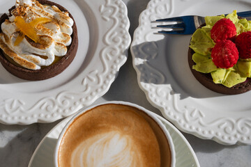a fresh cappuccino with latte art in a sage-green ribbed cup, placed in the foreground with two gourmet fruit tarts visible on white plates in the background.