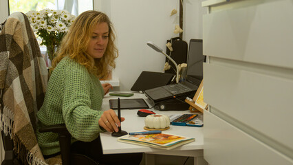 A woman in a green sweater uses a graphic tablet at a desk, surrounded by art supplies and a laptop.