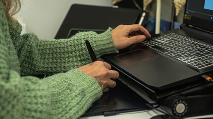 Close up of a hand using a stylus on a graphic tablet, connected to a laptop, in a creative workspace.