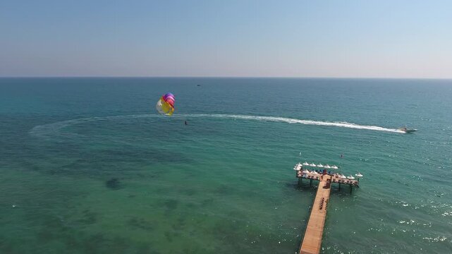 Aerial view of summer vacation tourists parasailing behind Revival boat near hotel pier. Adrenaline water sport scene shows colorful flight over sea during sunny holiday season.