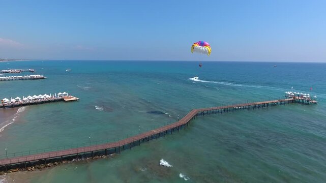 Aerial view of summer vacation tourists parasailing behind Revival boat near hotel pier. Adrenaline water sport scene shows colorful flight over sea during sunny holiday season.