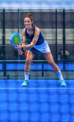 Smiling female padel player, ready to play, holding her racket on a blue court, behind the net.