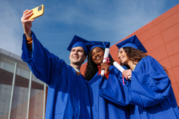 Diverse students in graduation caps and gowns holding diplomas, capturing a selfie