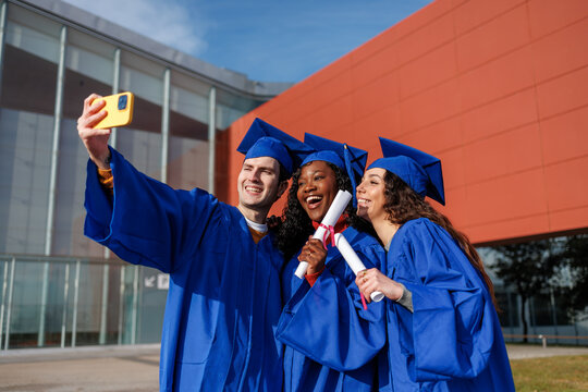 Happy diverse students celebrating graduation holding diplomas and taking a selfie
