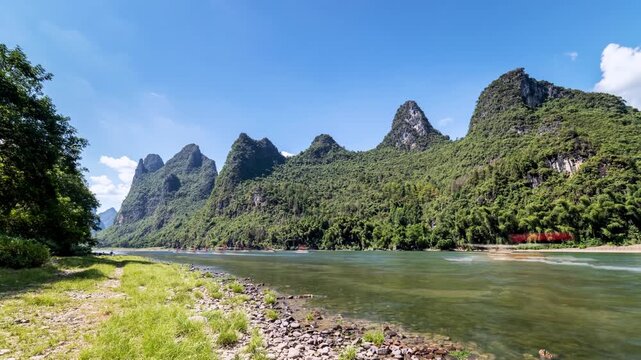slow-shutter time lapse on the Li River: hikers, boats, and bamboo rafts blur against karst peaks under blue skies in Yangshuo, China