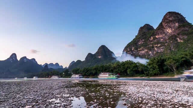slow-shutter timelapse of Li River at dusk, boats blur across pebbled banks and hearth smoke with karst peaks silhouetted under soft twilight in Yangshuo, Guilin, China