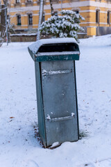 Snow covers a solitary trash bin beside a picturesque building in a serene winter park