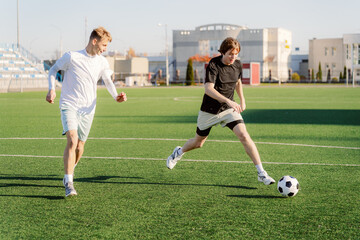 Two young men play soccer together on a vibrant green field, laughing and having fun as they kick the ball towards each other. The sun shines brightly, creating a joyful atmosphere.
