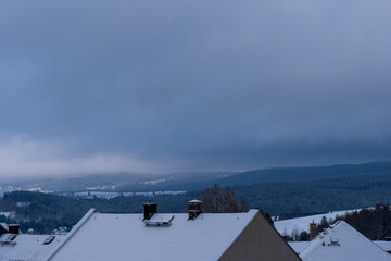 Snowy winter landscape captured from rooftops, showcasing a peaceful, cloudy sky and distant hills in soft hues