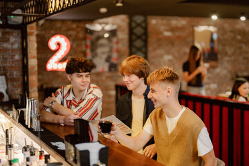 Three young men sit at the bar counter, chatting and laughing as they explore the drink menu. The cozy atmosphere features warm lights and a bustling vibe in the background.