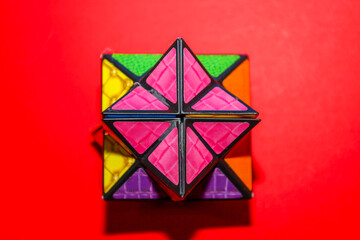 A complex multifaceted geometric puzzle with vibrant textured panels of pink, green, and purple colors set against a rich red background. This intellectual toy stimulates logical thinking,