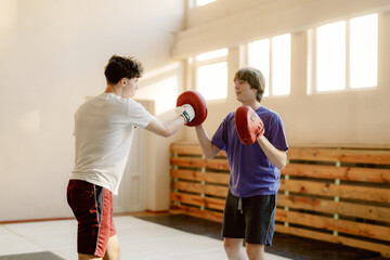 Two young men practice boxing techniques in a spacious gym with natural light. One wears gloves and strikes pads held up by the other, emphasizing teamwork and skill development.