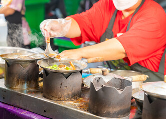 A street food vendor in a red shirt and face mask prepares a hot glass noodle casserole with shrimp