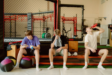 Three young men sit on mats in a gym, taking a break after an intense workout. They are enjoying drinks and checking their phones, surrounded by fitness equipment.