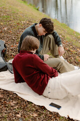 Fototapeta premium Two young men sit on a blanket near the water, sharing quiet moments and laughter. The autumn leaves create a warm atmosphere as they relax and enjoy each other's company.