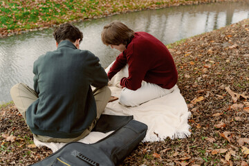 Two friends sit on a blanket by the river, surrounded by orange and yellow leaves. They are focused...