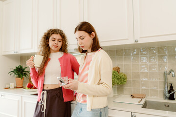 Two friends share a moment in a bright, welcoming kitchen. One sips coffee while the other scrolls...