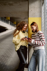 Two friends enjoy a light-hearted moment in a spacious parking garage. They share their phones...